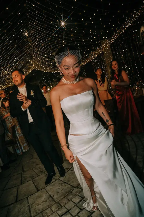 Bride in strapless satin corset split Aline look walking through the reception — birdcage veil and pearl necklace under warm string lights